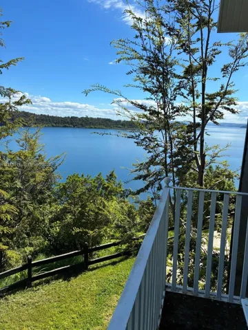 Elevated balcony with vertical railings overlooking a expansive lake, mature trees, and tiered grassy landscape featuring a post-and-rail fence