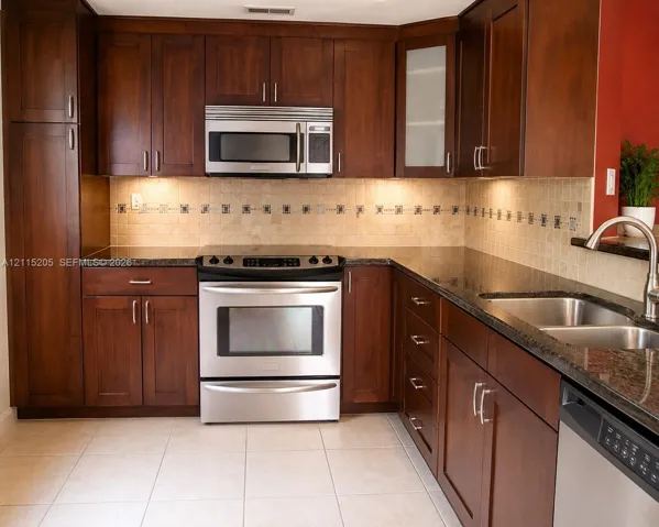Rich-toned cabinetry, stainless steel appliances, dark stone-finish countertops, tiled backsplash with decorative inlay, and under-cabinet lighting
