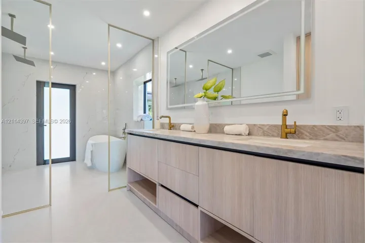 Dual-sink vanity with wood-finish cabinetry, natural stone countertop, brass fixtures, integrated LED-backlit mirror, and glass-enclosed wet room featuring a soaking tub