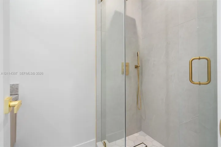 Walk-in shower featuring large-format grey tile walls, white hexagonal floor tile, brass-finish handheld faucet, and a frameless glass door with a coordinated brass handle