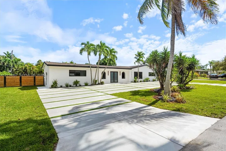 Single-story residence featuring a smooth stucco exterior, expansive concrete driveway with grass inlays, arched entryway, horizontal wood-slat privacy fencing, and mature palm landscaping
