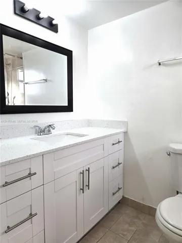 White shaker-style cabinetry with satin nickel pulls, stone-composite countertop with integrated sink, rectangular mirror with a black frame, three-light vanity fixture, and neutral tile flooring
