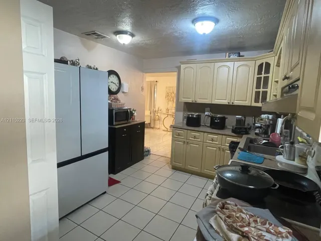 Kitchen featuring cream-toned cabinetry with glass-panel accents, solid surface countertops, a stainless steel multi-door refrigerator, tile flooring, and dual flush-mount ceiling light fixtures