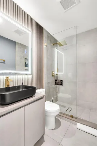 Bathroom featuring a slate-toned vessel sink, gold-tone hardware, a wall-mounted LED-backlit mirror, vertical slat accent wall, and a glass-enclosed walk-in shower with large-format tile