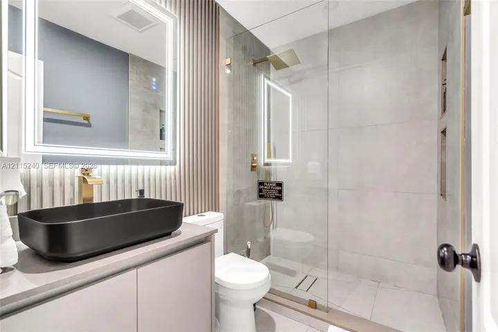 Bathroom featuring a matte black vessel sink, gold-toned faucet hardware, wall-mounted LED-backlit mirror, vertical slat accent wall, and a walk-in shower with large-format tile and a glass enclosure