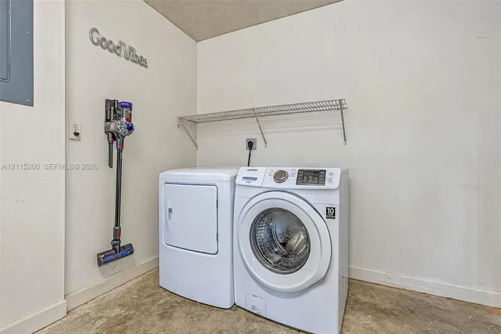 Laundry area featuring white washer and dryer units, a wall-mounted wire shelf, and exposed concrete flooring