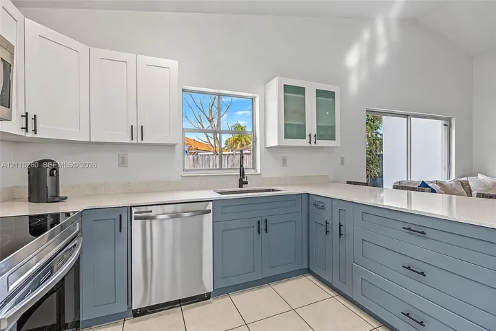 Kitchen featuring two-tone cabinetry with matte black hardware, stone-composite countertops, stainless steel dishwasher, glass-front upper cabinets, and tile flooring