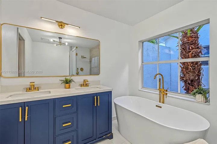 Dual-sink vanity with navy blue cabinetry, gold-tone hardware, and a quartz countertop, paired with a freestanding soaking tub featuring a gold-tone floor-mounted faucet and an expansive gold-rimmed mirror