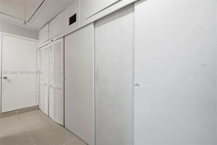 Wall-to-wall built-in storage featuring floor-to-ceiling smooth-panel sliding doors, overhead cabinet compartments, and louvered-style closet doors, complemented by ceramic tile flooring