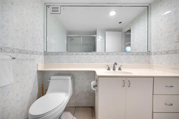 Bathroom featuring a single-basin vanity with a solid-surface countertop, floor-to-ceiling tiled walls, an expansive wall-mounted mirror, and a low-profile toilet