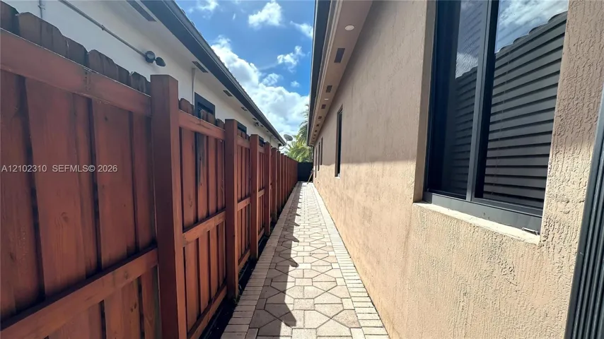 Stucco exterior wall featuring dark-framed windows and a stone-paver side path bordered by a wood privacy fence