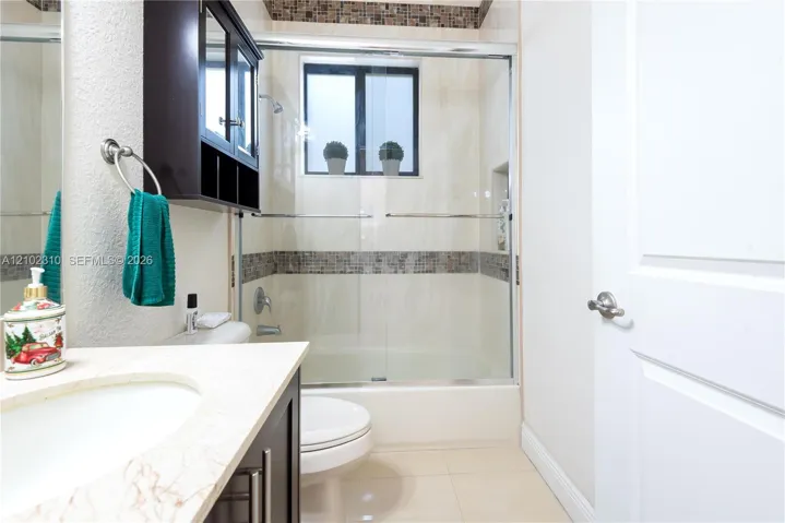 Bathroom featuring a dark-finish vanity with a stone-top integrated sink, a tiled shower-tub combination with sliding glass doors and decorative mosaic inlay, and ceramic tile flooring