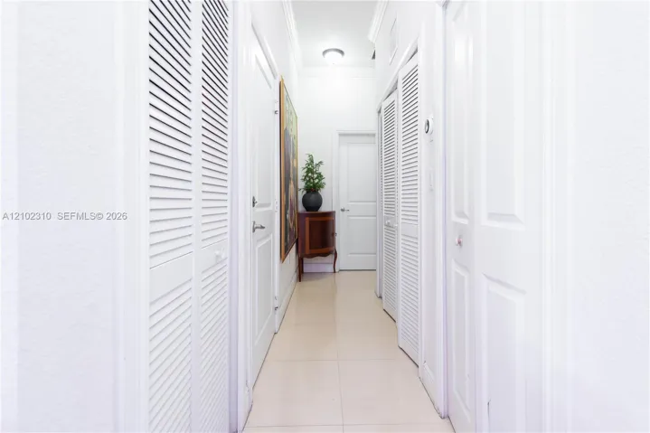 Hallway featuring neutral large-format tile flooring, multiple sets of white louvered-style closet doors, solid-panel interior doors, and crown molding