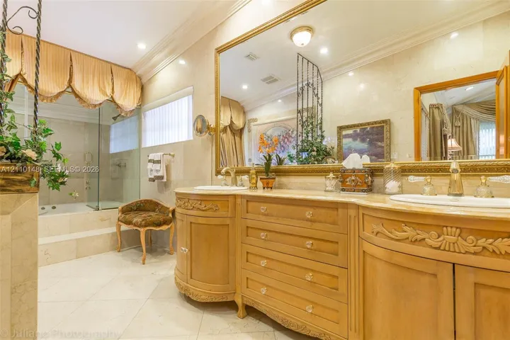 Dual-vanity cabinetry with intricate wood carvings and stone countertops, large wall-mounted mirror with gold-tone detailing, stone wall and floor tile, and a glass-enclosed jetted tub