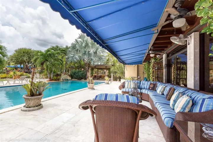 Covered outdoor patio with a retractable blue awning, stone-paver decking, rectangular swimming pool, and ceiling fans