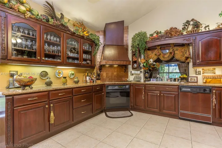 Custom dark wood cabinetry with glass-front display uppers, expansive stone-surface countertops, integrated range hood, built-in oven, and ceramic tile flooring