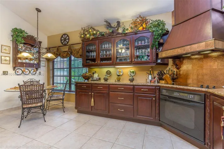 Kitchen featuring custom wood cabinetry with glass-front display uppers, stone-surface countertops, decorative metallic backsplash, a large wood-finish range hood, and tile flooring