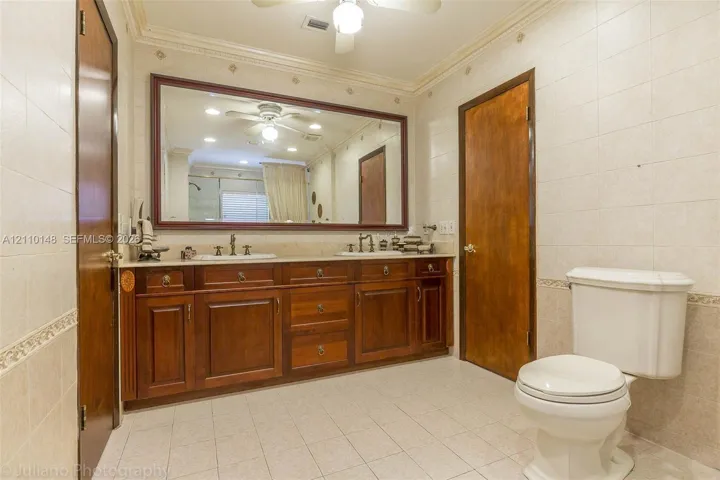 Dual-sink vanity with wood-finish cabinetry, solid surface countertop, expansive wall-mounted mirror, and tile-finished walls