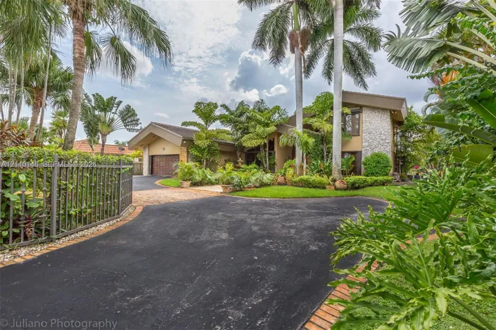 Asphalt driveway with brick-paver accents, stone exterior siding, multi-gabled roofline, metal perimeter fencing, and mature tropical landscaping