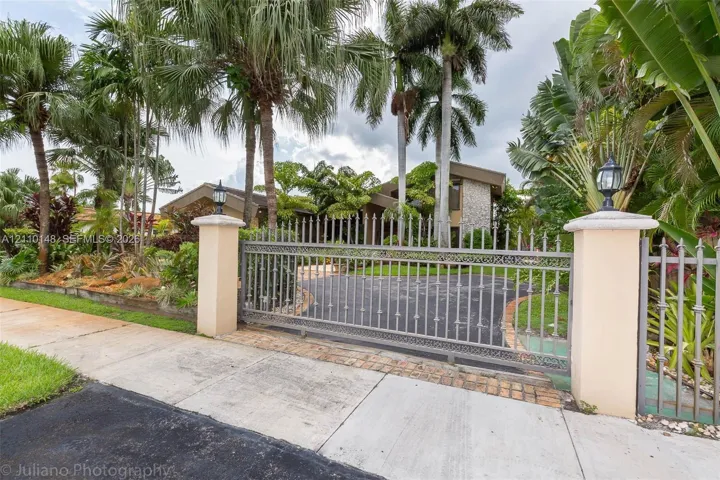 Gated entrance featuring decorative metal fencing, stone-topped stucco pillars with mounted lighting, and a paved driveway surrounded by mature tropical landscaping