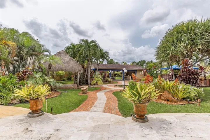 Landscaped backyard featuring a stone-paved patio, a thatched-roof gazebo, mature tropical vegetation, and a brick walkway