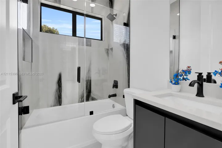 Bathroom featuring a dark-toned floating vanity with a white countertop, large-format tile surround, frameless glass shower panel, and matte black fixtures