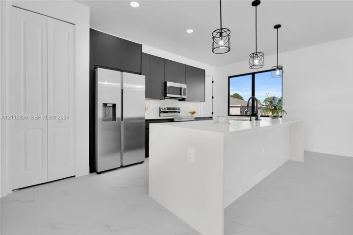 Kitchen featuring flat-panel dark cabinetry, white countertops, stainless steel appliances, a central island with an integrated sink, and pendant lighting
