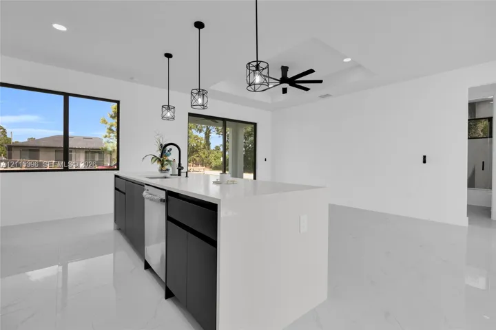 Kitchen island with waterfall-edge countertop, dark cabinetry, recessed ceiling with black ceiling fan, black metal pendant lighting, and polished stone-finish flooring