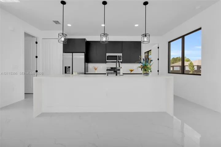 Kitchen featuring a large central island with a solid-surface countertop, flat-panel dark cabinetry, stainless steel appliances, pendant lighting, and polished stone-finish flooring