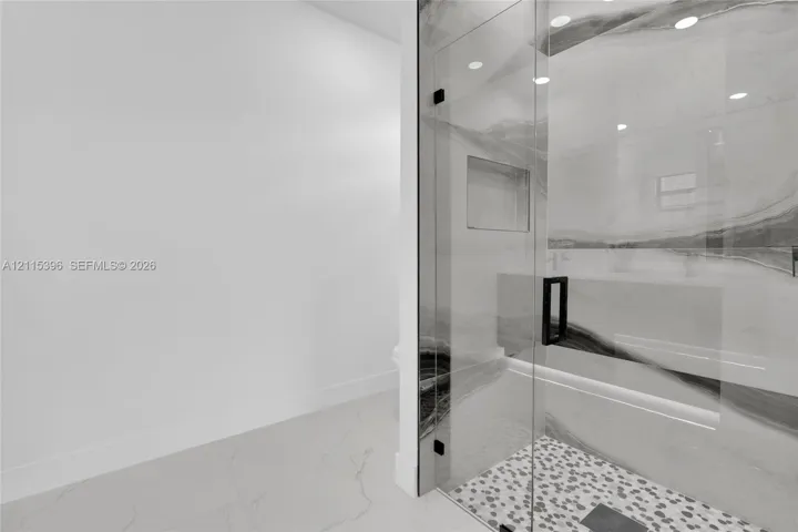 Walk-in shower featuring large-format patterned wall tile, a frameless glass door with matte black hardware, integrated recessed wall niche, pebble-textured floor tile, and recessed ceiling lighting