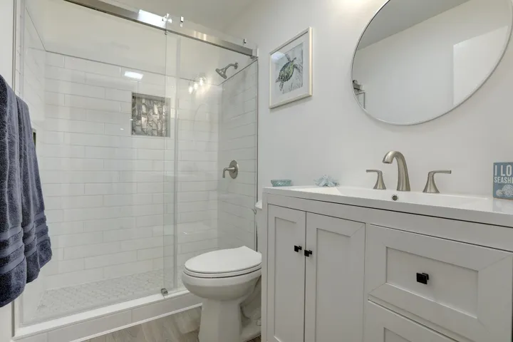 Bathroom featuring a white vanity cabinet with a rectangular basin, oval wall-mounted mirror, walk-in shower with white subway tile surround, glass enclosure, and wood-finish flooring