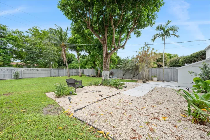 Fenced backyard featuring a manicured lawn, stone hardscaping, mature trees, and palm vegetation