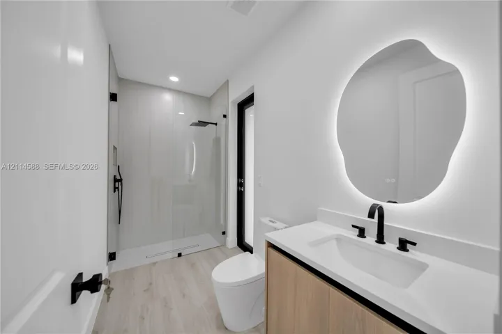 Bathroom featuring a wood-finish vanity, white countertop, illuminated organic-shaped mirror, matte black fixtures, and a walk-in glass-enclosed shower