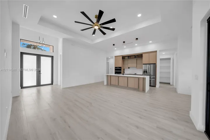 Tray ceiling with integrated recessed lighting, light wood-finish flooring, dual French entry doors with transom window, kitchen featuring light wood cabinetry, and waterfall-edge island