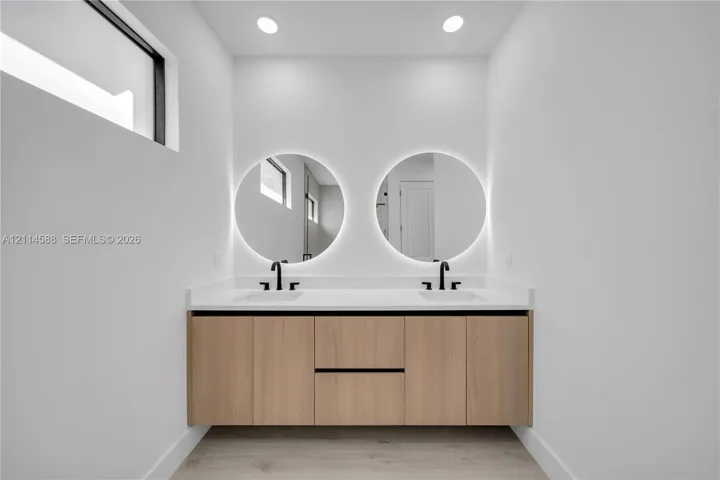Floating vanity with light wood-finish cabinetry, dual rectangular undermount sinks, quartz countertop, matte black widespread faucets, and dual circular backlit mirrors