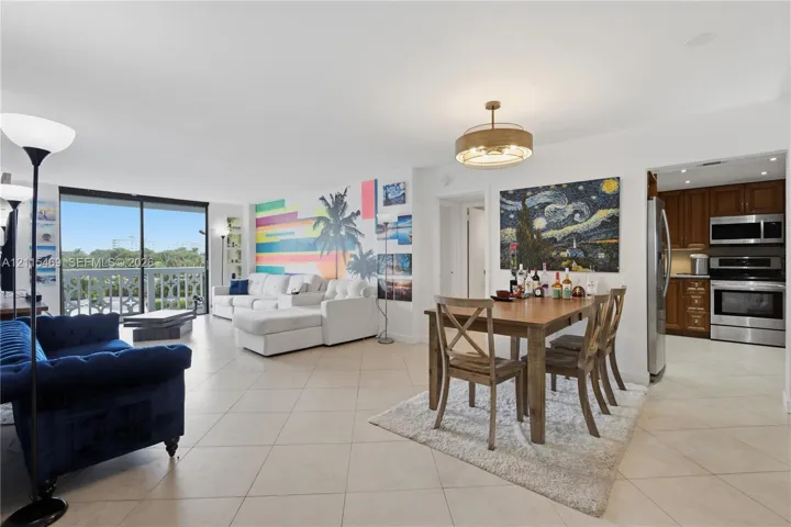 Open-concept living area featuring light-toned tile flooring, large sliding glass doors, a contemporary drum chandelier, and a kitchen equipped with dark wood-finish cabinetry and stainless steel appliances