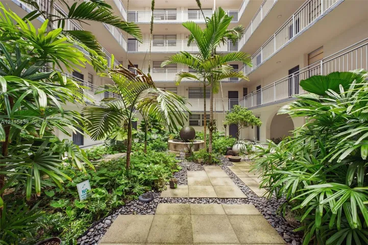 Central landscaped atrium featuring stone paving, a tiered spherical water feature, mature palm trees, and multi-level balconies with metal railings
