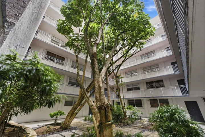 Open-air central courtyard featuring mature trees, a stone paver walkway, multi-level balconies, and a neutral stucco facade