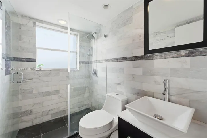 Full-height shower enclosure with glass door, stone-tile wall surround, vessel sink vanity, contrasting dark-framed mirror, and integrated recessed lighting