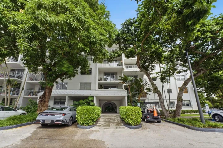 Multi-story residential building exterior featuring private balconies, a covered ground-level entryway, and mature shade trees