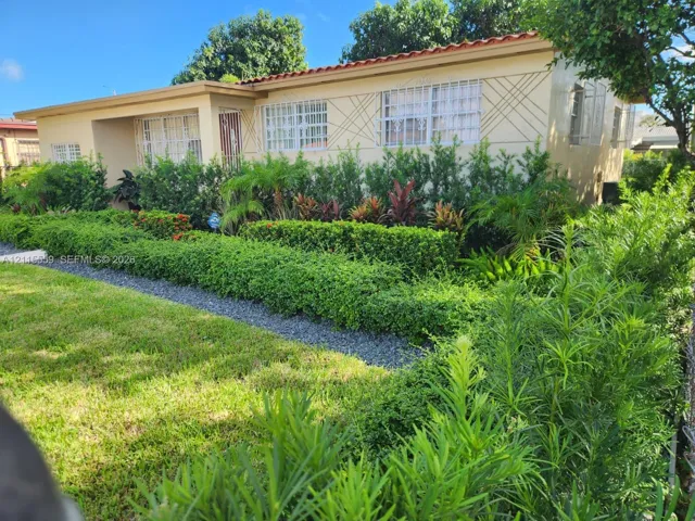 Single-story exterior featuring a decorative stucco facade, tiled roof, geometric wall accents, metal window security bars, and extensive landscaped shrubbery