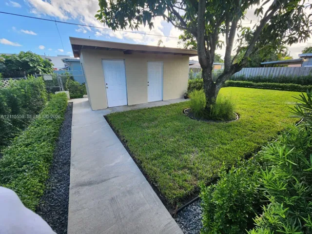 Detached structure featuring dual exterior doors, concrete walkway, manicured lawn, and perimeter landscaping