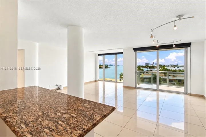 Open-concept interior featuring large-format tile flooring, a stone-surface breakfast bar, structural pillar, adjustable track lighting, and floor-to-ceiling glass sliding doors opening to an exterior balcony
