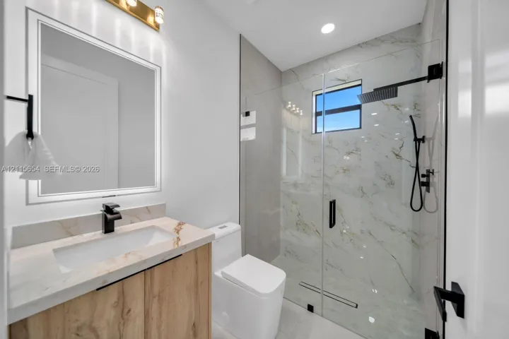 Bathroom featuring a wood-finish vanity with a rectangular sink, stone-surface countertop, frameless glass-enclosed shower with wall-mounted rain showerhead, and matte black fixtures