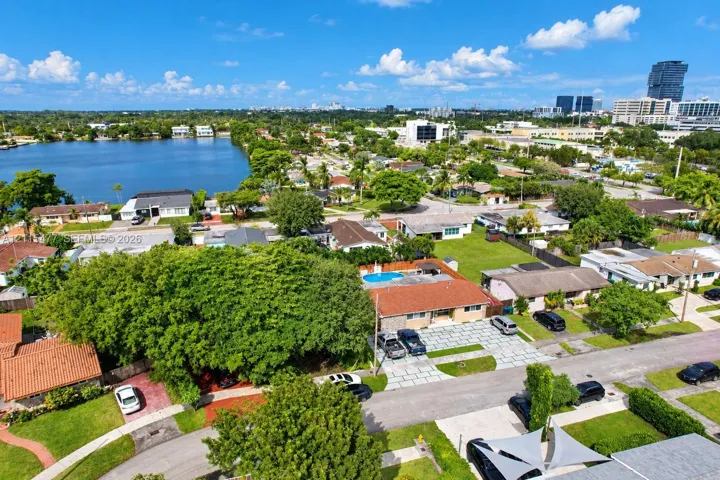 Aerial perspective featuring a single-story residence with a red-tiled roof, an inground swimming pool, a multi-vehicle paved driveway, and mature trees situated within an urban residential setting