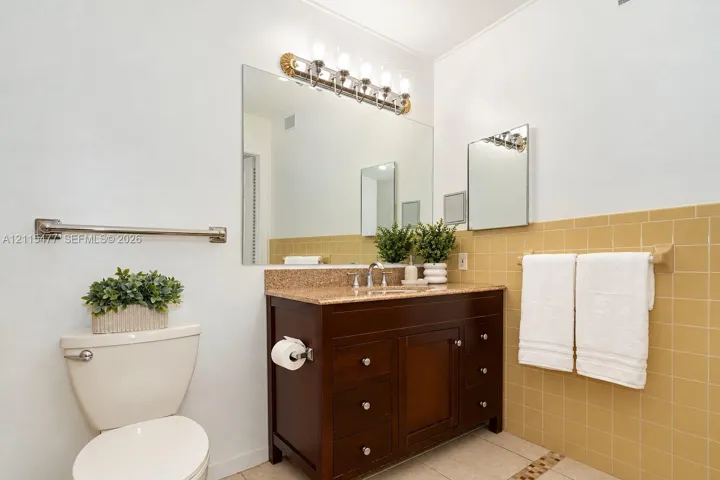 Bathroom featuring a dark wood-finish vanity, stone-surfaced countertop with an integrated basin, wall-to-wall mirror, multi-light fixture, and ceramic tile wall accents