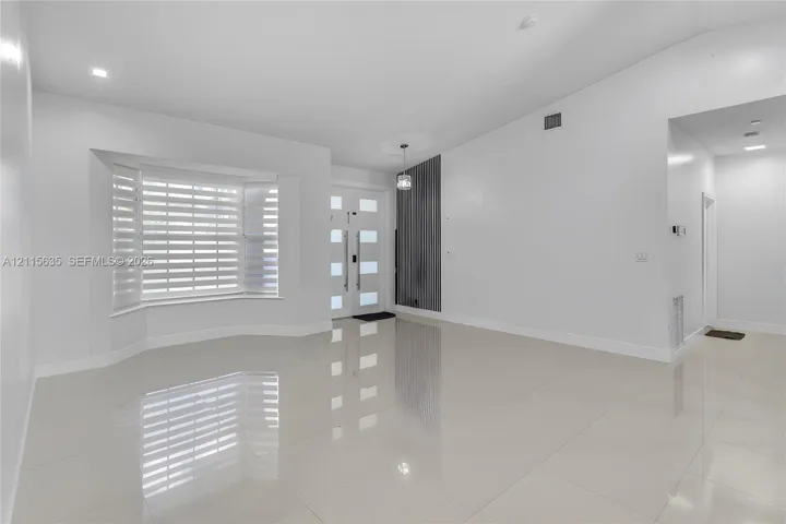 Bright interior featuring vaulted ceilings, polished tile flooring, a multi-pane front entry door with vertical handle hardware, and a bay window equipped with horizontal blinds