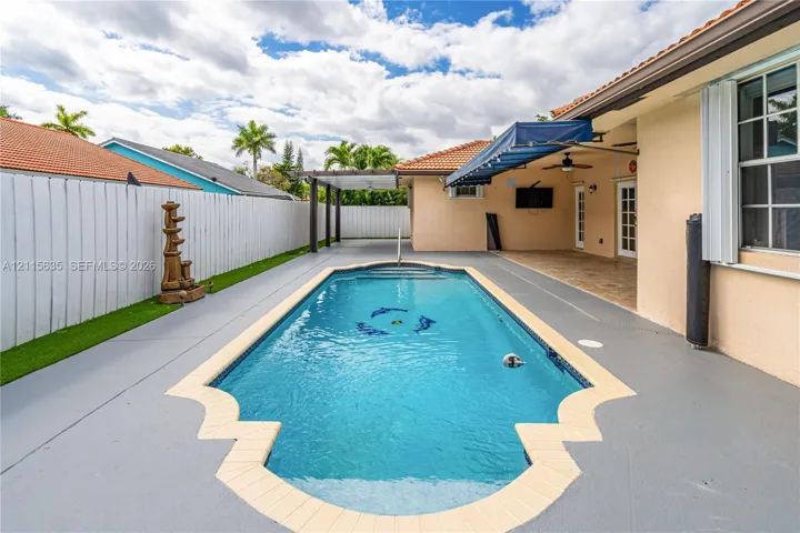 In-ground swimming pool with decorative tile coping, covered patio, retractable fabric awning, and enclosed outdoor area with white privacy fencing
