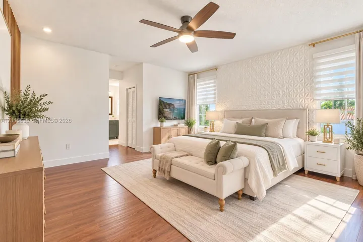 Bedroom featuring wood-finish flooring, a textured geometric accent wall, a central ceiling fan with integrated lighting, and dual windows with layered window treatments