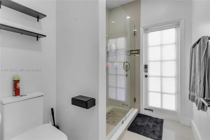 Full-height glass-enclosed shower, wall-mounted matte black shelving, matte black wall-mounted toilet paper holder, and a glass-paneled exterior door